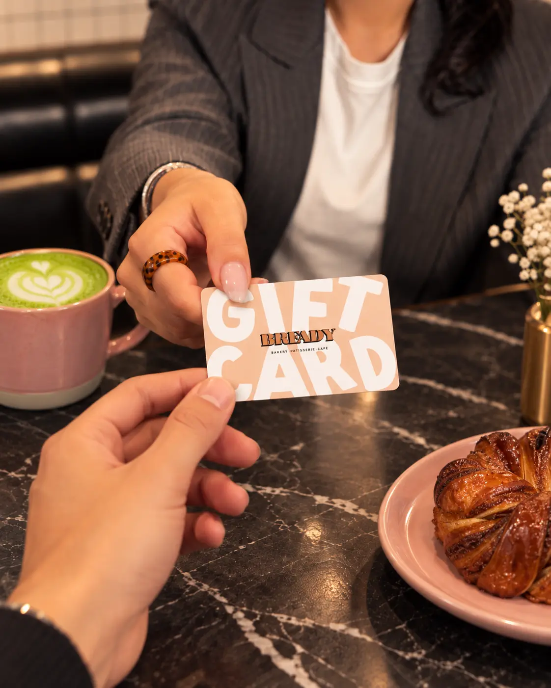 A hand passing a Bready gift card across a café table beside a latte and a pastry