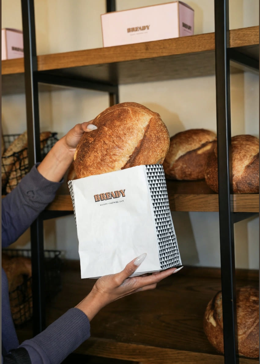 A hand holding a Bready paper bag with a fresh sourdough loaf, in front of shelves of bread