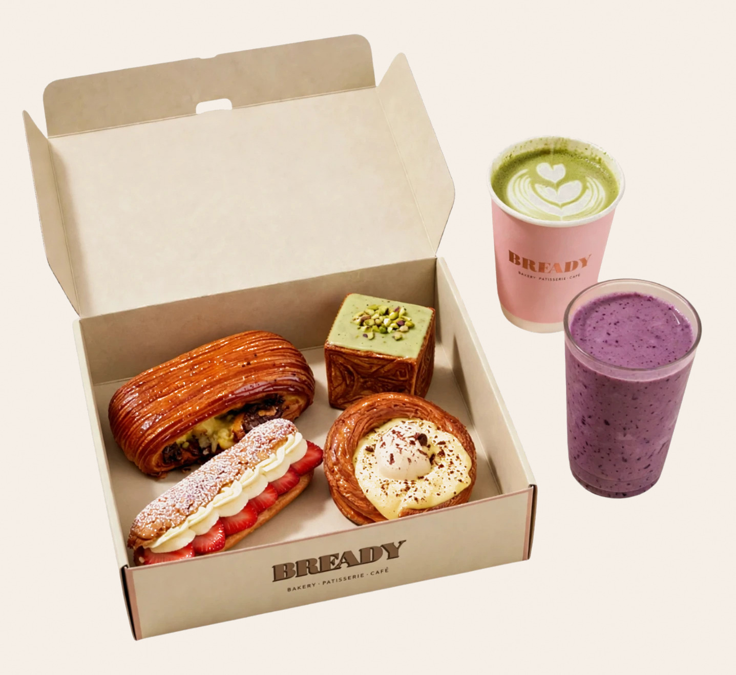 An open Bready box holding four pastries — a chocolate roll, a pistachio cube loaf, a strawberry cream pastry and a danish — beside a matcha latte in a pink Bready cup and a berry smoothie